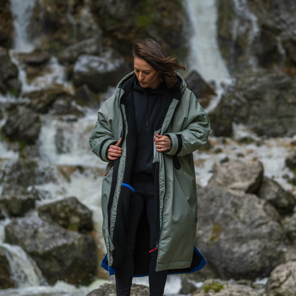Woman wearing a green coat standing in front of a waterfall and rocks.
