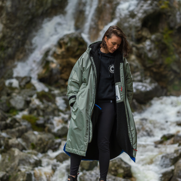Person wearing a green coat walking on rocky path with a waterfall in the background